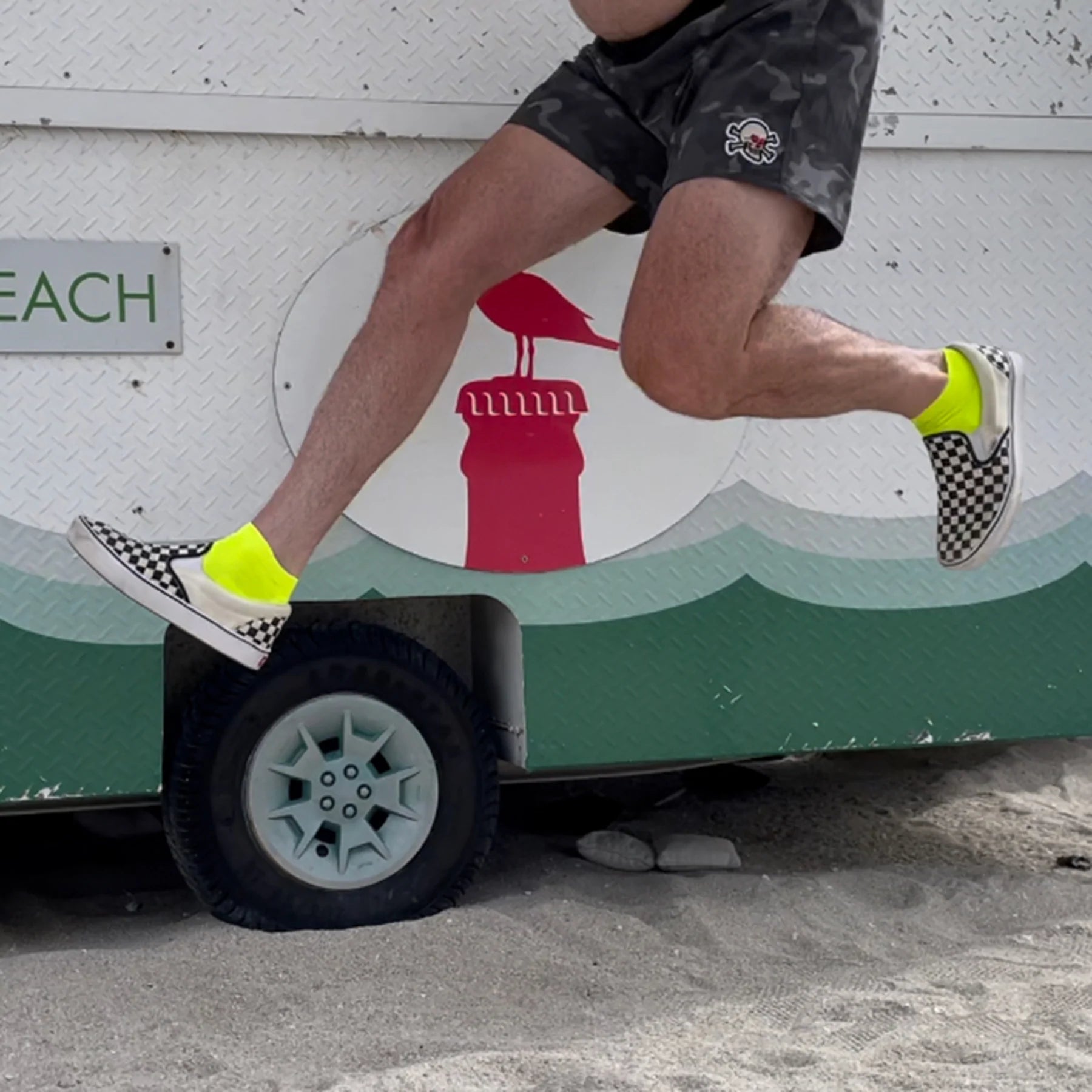 man jumping on beach in front of van wearing checkerboard Vans and bright yellow DeFeet All Day cycling socks with a 1" cuff