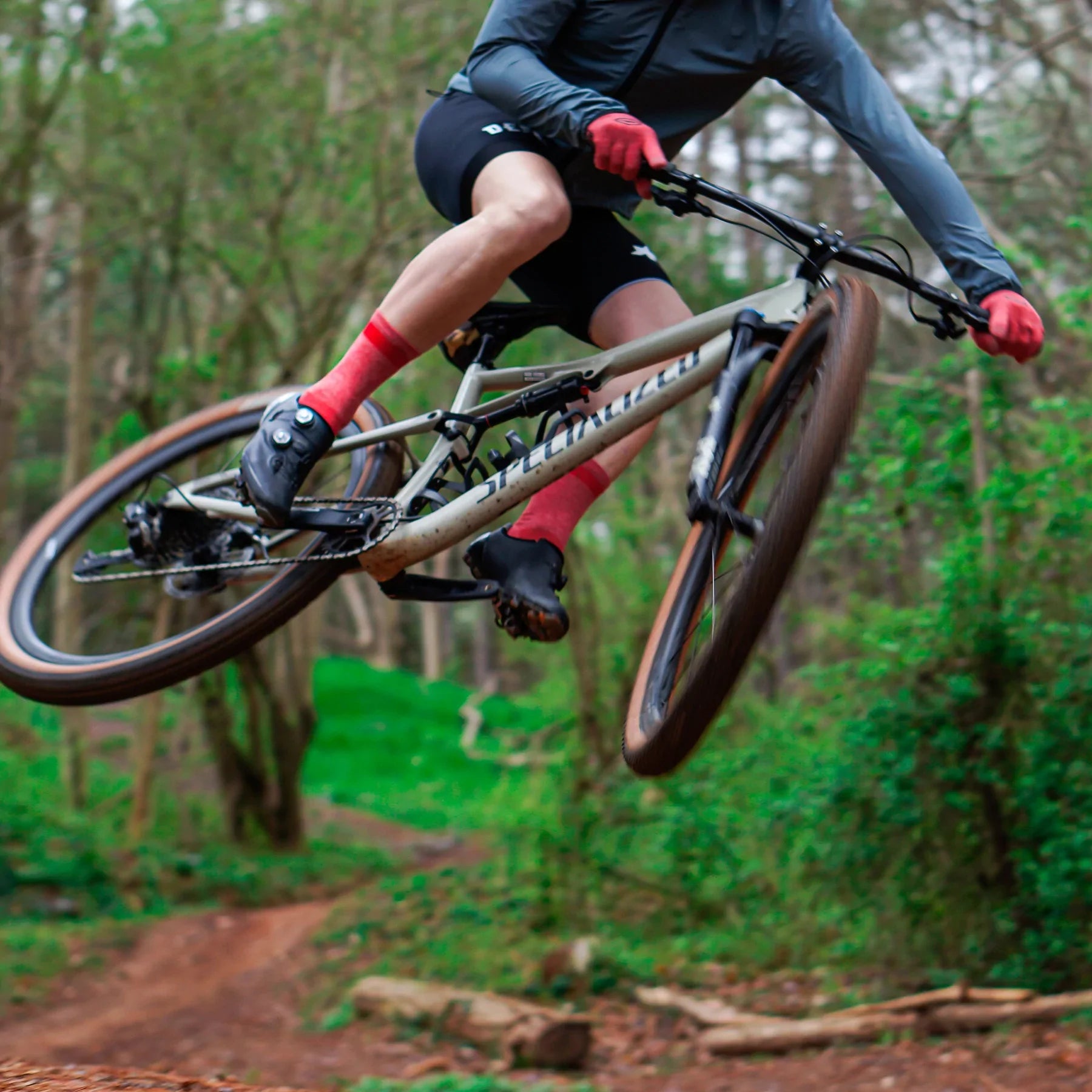 white male cyclist jumping a bike on a wooded trail, wearing red gloves and red cycling socks